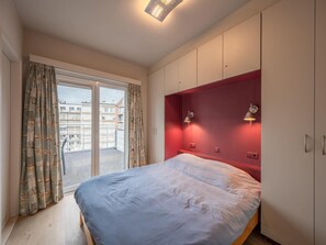 A bedroom with a bed, a red accent wall, a sliding glass door, and a view of buildings outside. - Stylish Stay by Rubensplein (Knokke-Heist)
