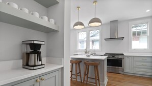 Una cocina moderna con armarios blancos, una cafetera, taburetes de madera, luces colgantes y un ventanal grande.