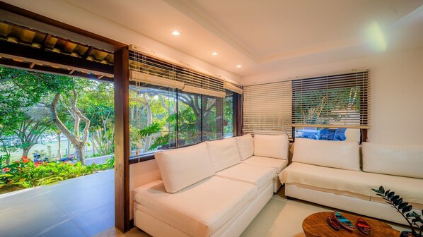 A modern living room with a white sofa, large windows with blinds, and a view of greenery outside.