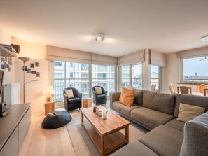 A modern living room with a sofa, chairs, a coffee table, and a view of buildings through large windows. - Apartment in Knokke Near the Beach (Knokke-Heist)