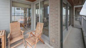 A covered outdoor area with wooden chairs and a table, a glass door leading inside, and a brick pillar. - Apartment in Knokke Near the Beach (Knokke-Heist)