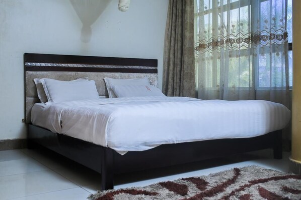 A bed with a dark headboard, white bedding, and a patterned rug on a tiled floor. - Excellent Hotel Kampala (Kampala)