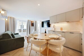 Uma sala de estar moderna com um sofá e uma mesa de jantar com cadeiras. Há uma área de cozinha com armários e uma TV fixada na parede.