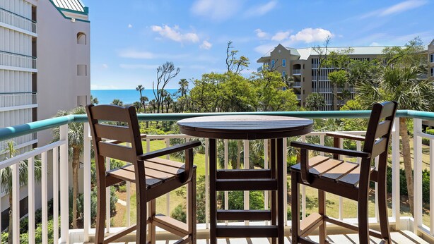 A balcony with two wooden chairs and a small round table, overlooking a view of the ocean and a resort area with buildings and palm trees.