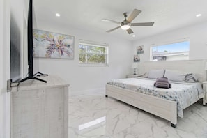 A bedroom with a bed, a ceiling fan, a TV on a stand, a window, and artwork on the walls.