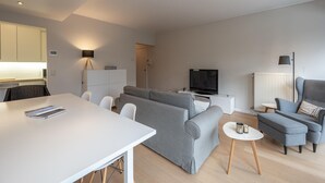A modern living room with a grey sofa, a white dining table, and a flat-screen TV. - Apartment in Knokke Near Duinbergen Beach (Knokke-Heist)