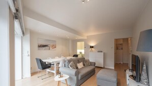 A modern living room with a dining area, grey sofas, a white dining table, and a flat-screen TV. - Apartment in Knokke Near Duinbergen Beach (Knokke-Heist)