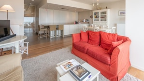 Ein modernes Wohnzimmer mit einem roten Sofa, einem Couchtisch mit Zeitschriften, einem Fernseher und einer Küchenzeile im Hintergrund.