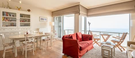 A spacious living area with a red sofa, wooden chairs, a dining table, and a view of the ocean through large windows.