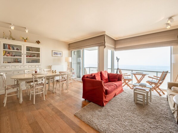 Amplia sala de estar con un sofá rojo, sillas de madera, una mesa de comedor y vista al océano a través de grandes ventanales.