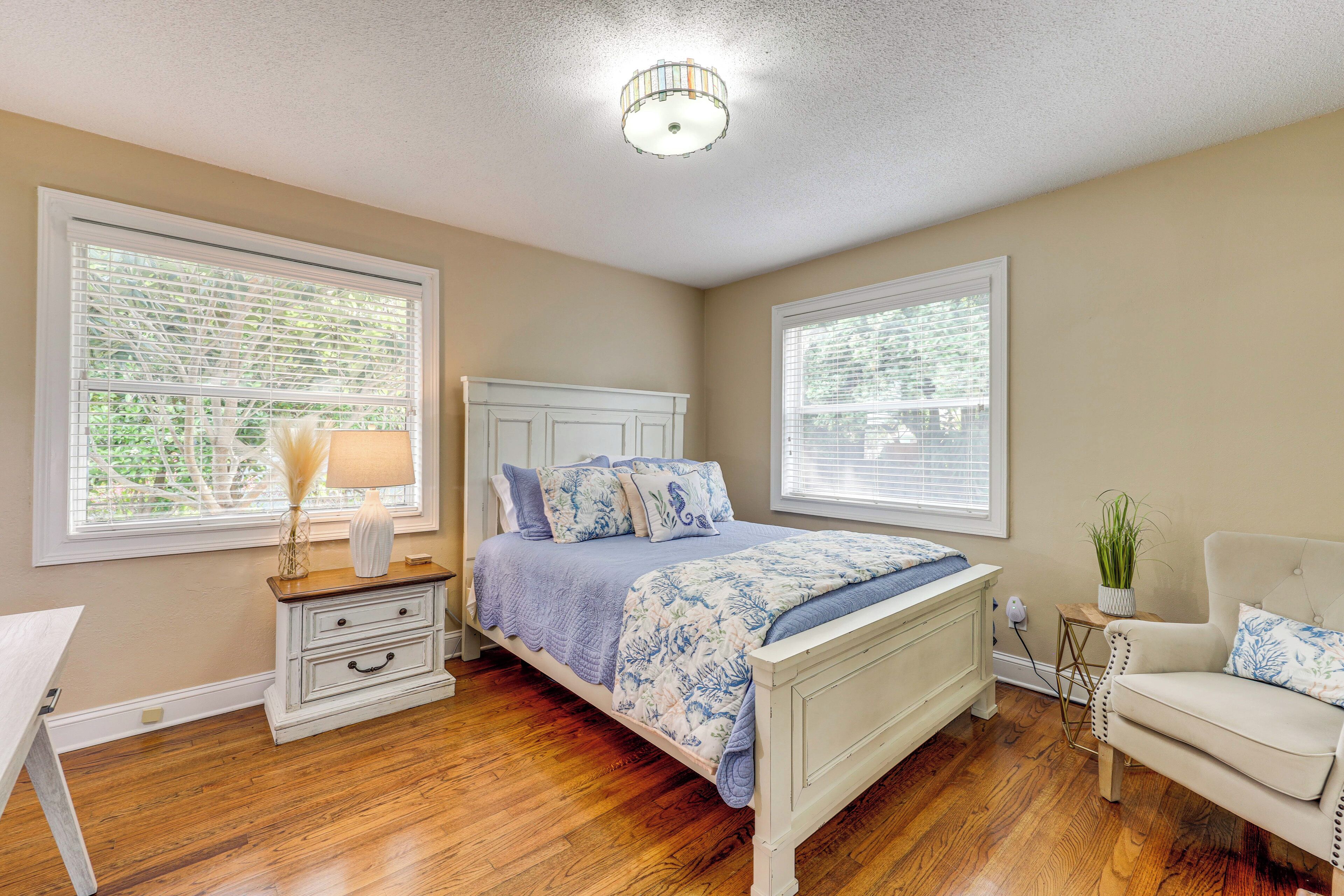 A bedroom with a white bed, a nightstand, a chair, and two windows with blinds.
