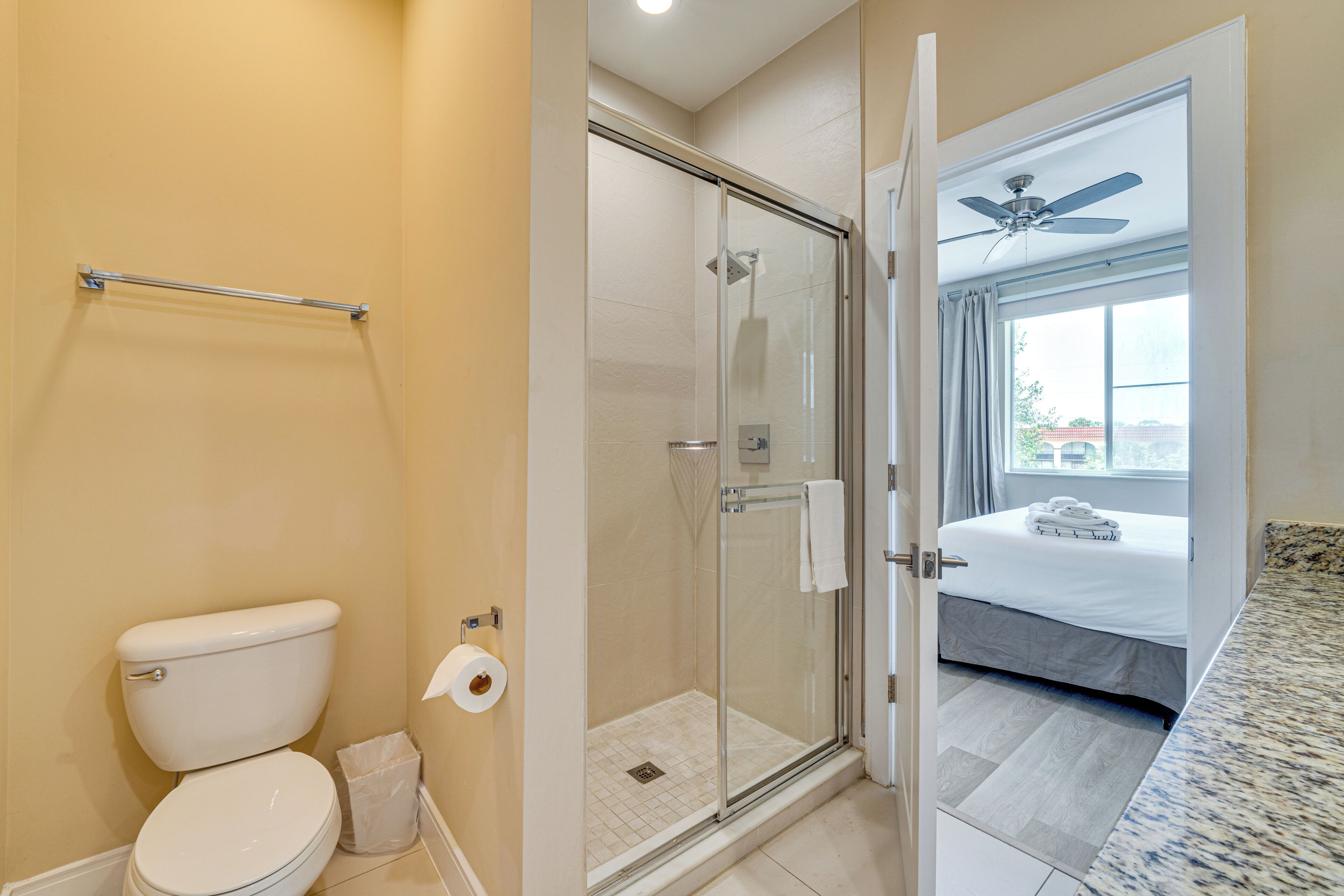 A bathroom with a toilet, a glass shower enclosure, and a bed in the adjacent room.