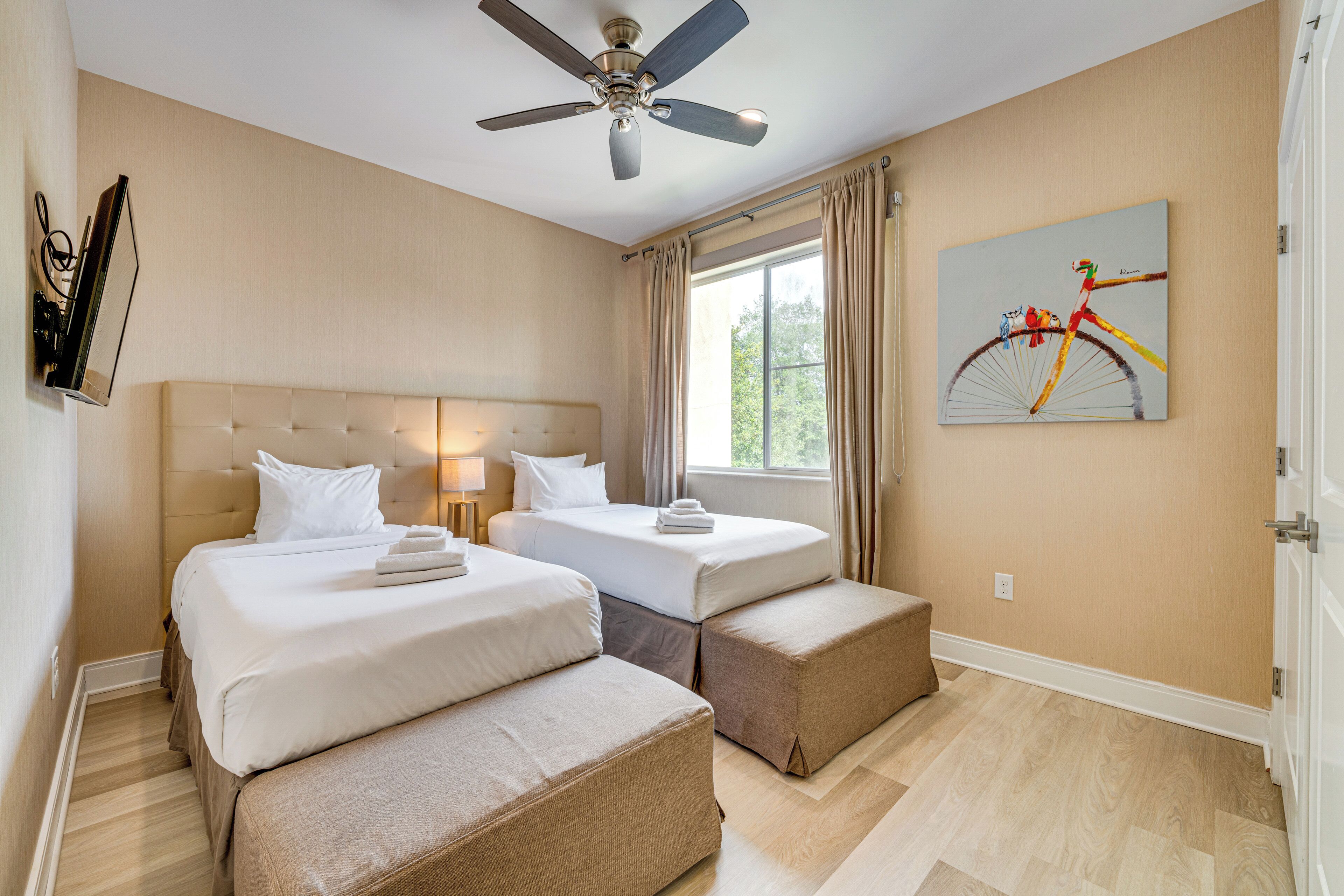 Quarto de hotel com duas camas, ventoinha no teto, janela com cortinas e um quadro na parede.