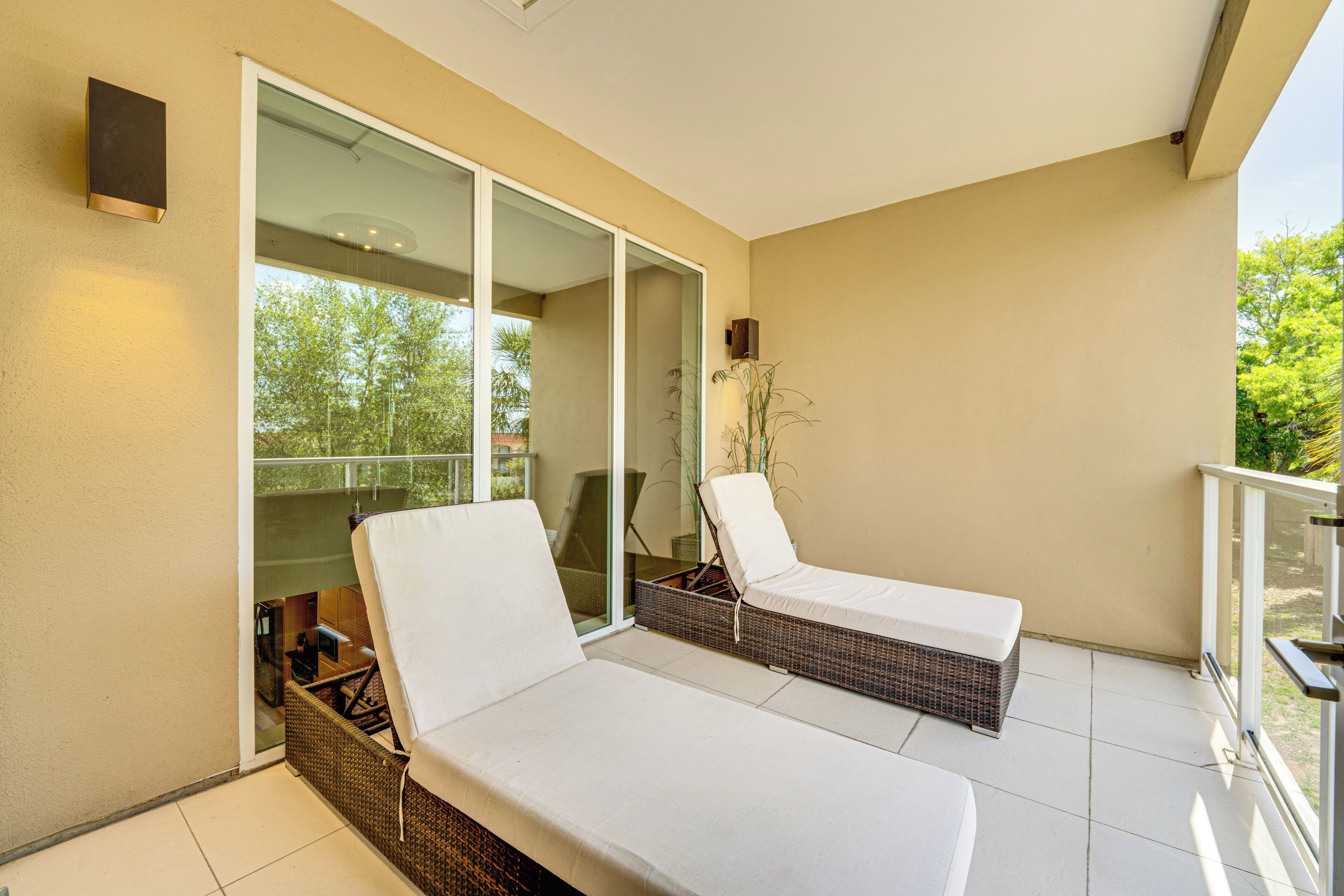 Un balcón con dos sillones blancos acolchados y un banco de mimbre, una puerta de cristal que da a un patio, y una pequeña mesa con una planta.