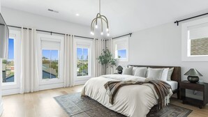 A bedroom with a large bed, a nightstand, a lamp, and a view of the city through large windows. - The Blackstone Manor (Atlanta)