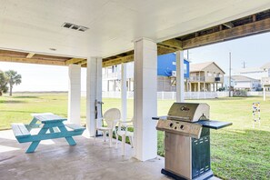A covered patio area with a picnic table and grill, surrounded by white chairs and a view of houses in the background. - Walk to Beach: Galveston Home w/ Patio & Balcony! (Galveston)