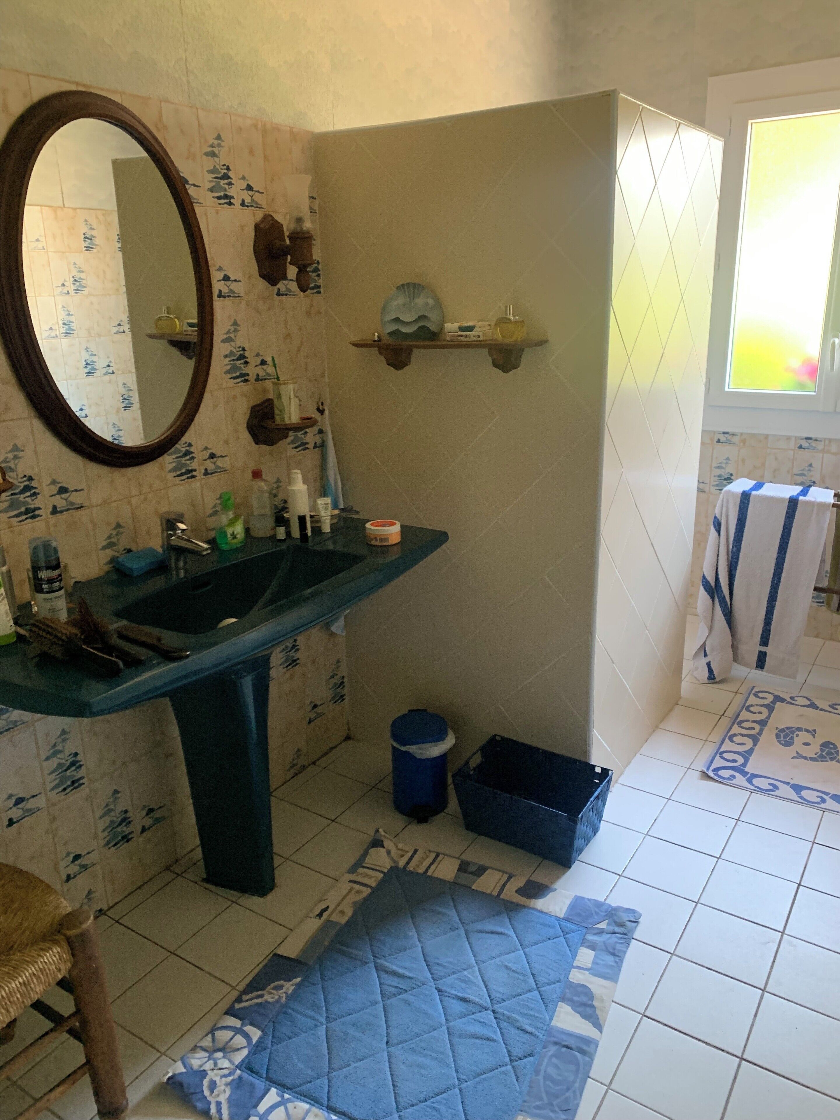 A bathroom with a sink, mirror, and tiled floor. There is a blue trash bin and a blue mat on the floor.