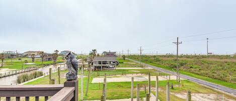 A view from a wooden deck overlooking a coastal area with a building, grassy fields, and a road.