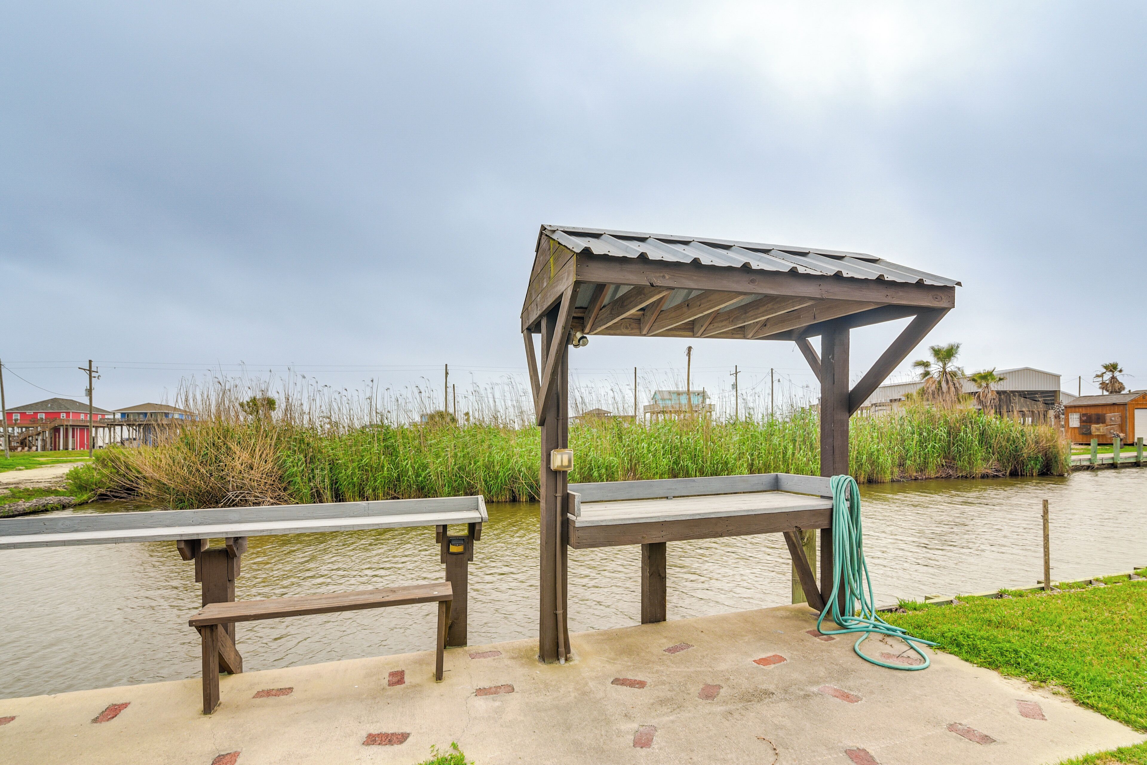 Un kiosque en bois avec des bancs, un tuyau d’arrosage et un chemin en béton, le tout situé près d’un plan d’eau.