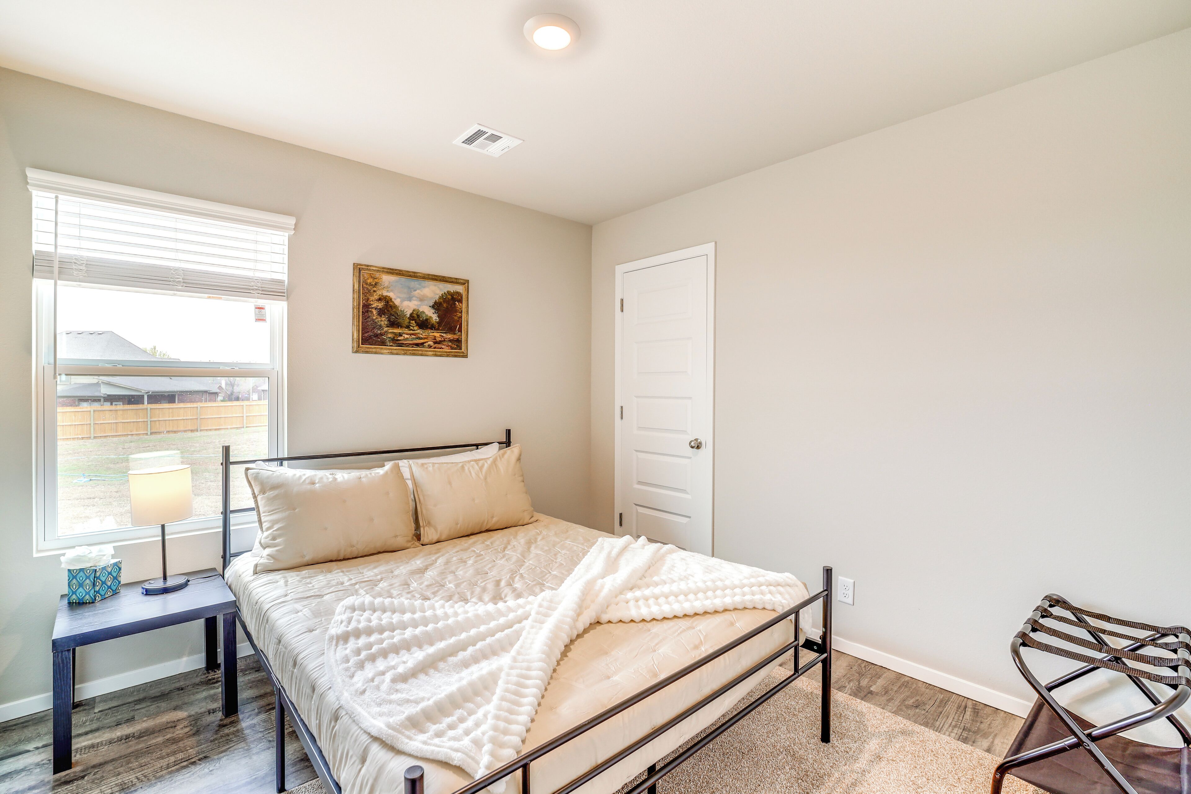 A bedroom with a bed, a nightstand, a chair, a window with blinds, a door, and a painting on the wall.