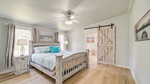A bedroom with a wooden bed, a ceiling fan, a window with curtains, and a door with a barn-style design. - Walk to Beach: Eufaula 'fishermans Delight' Home (Eufaula)