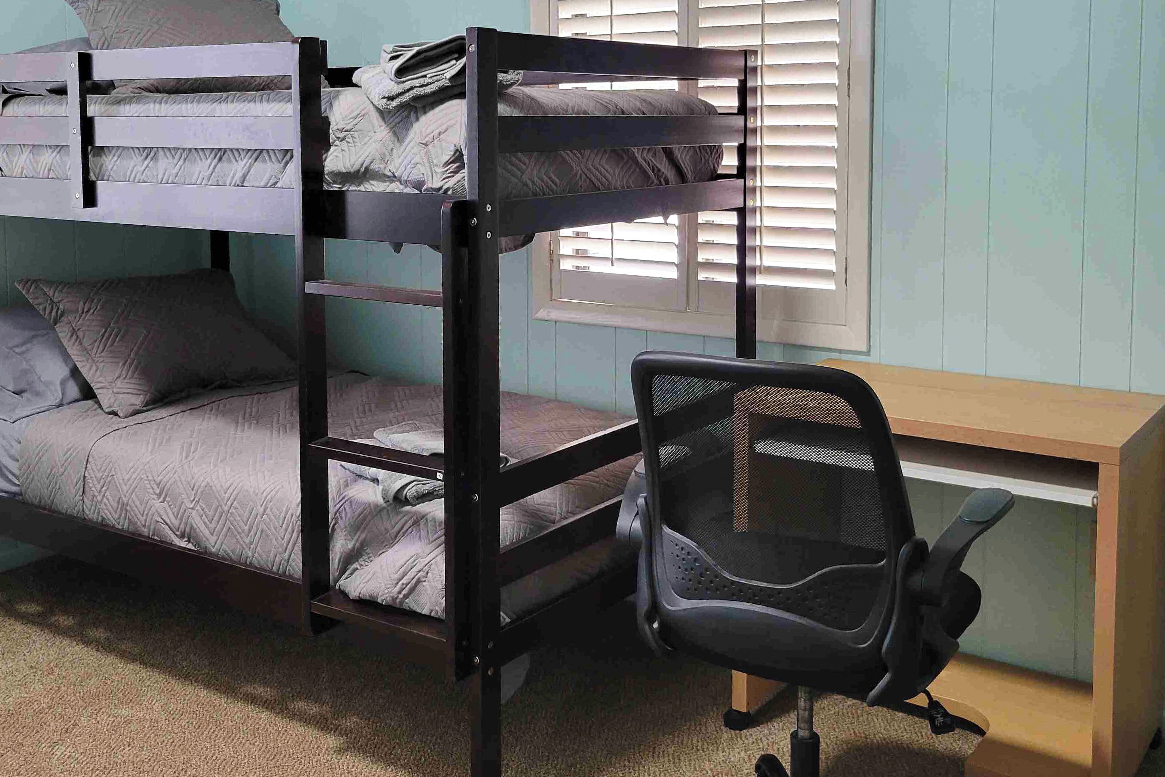 A bunk bed with grey bedding, a black office chair, and a wooden desk in a room with light blue walls and a window with blinds.