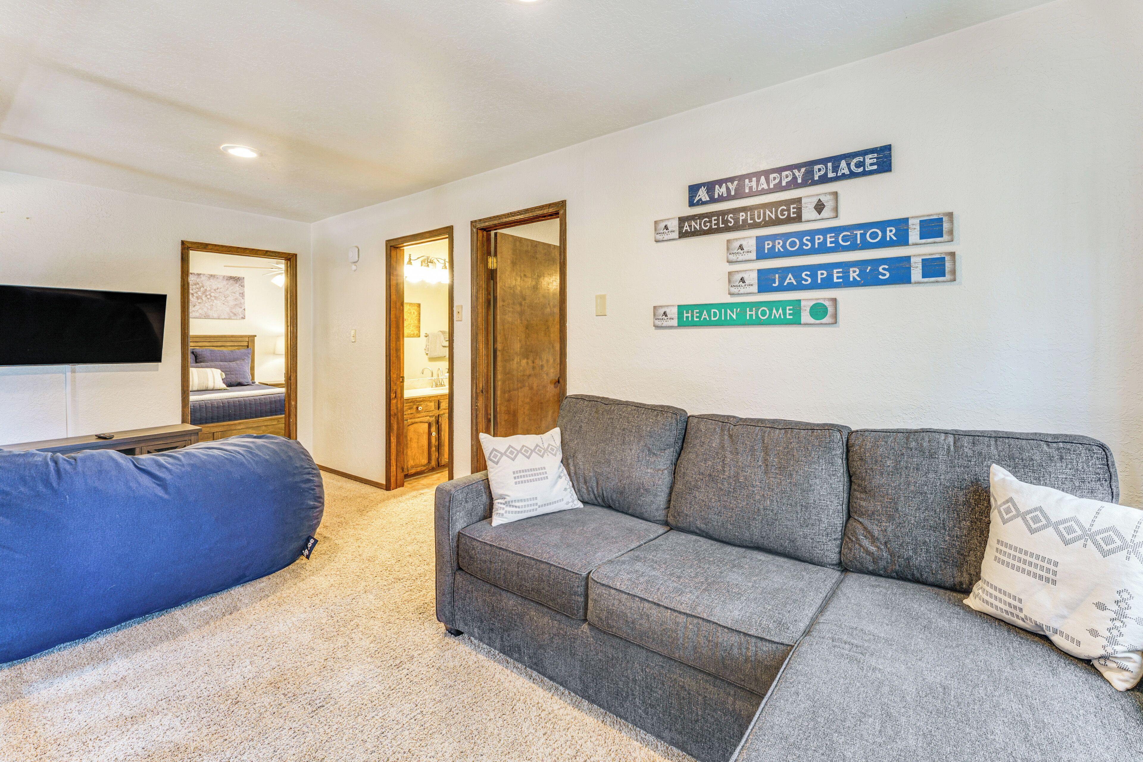 A living room with a grey sofa, a blue bean bag, and a wall with signs reading "MY HAPPY PLACE," "ANGEL'S PLUGNE," "PROSPECTOR," "JASPER'S," and "HEADIN' HOME."