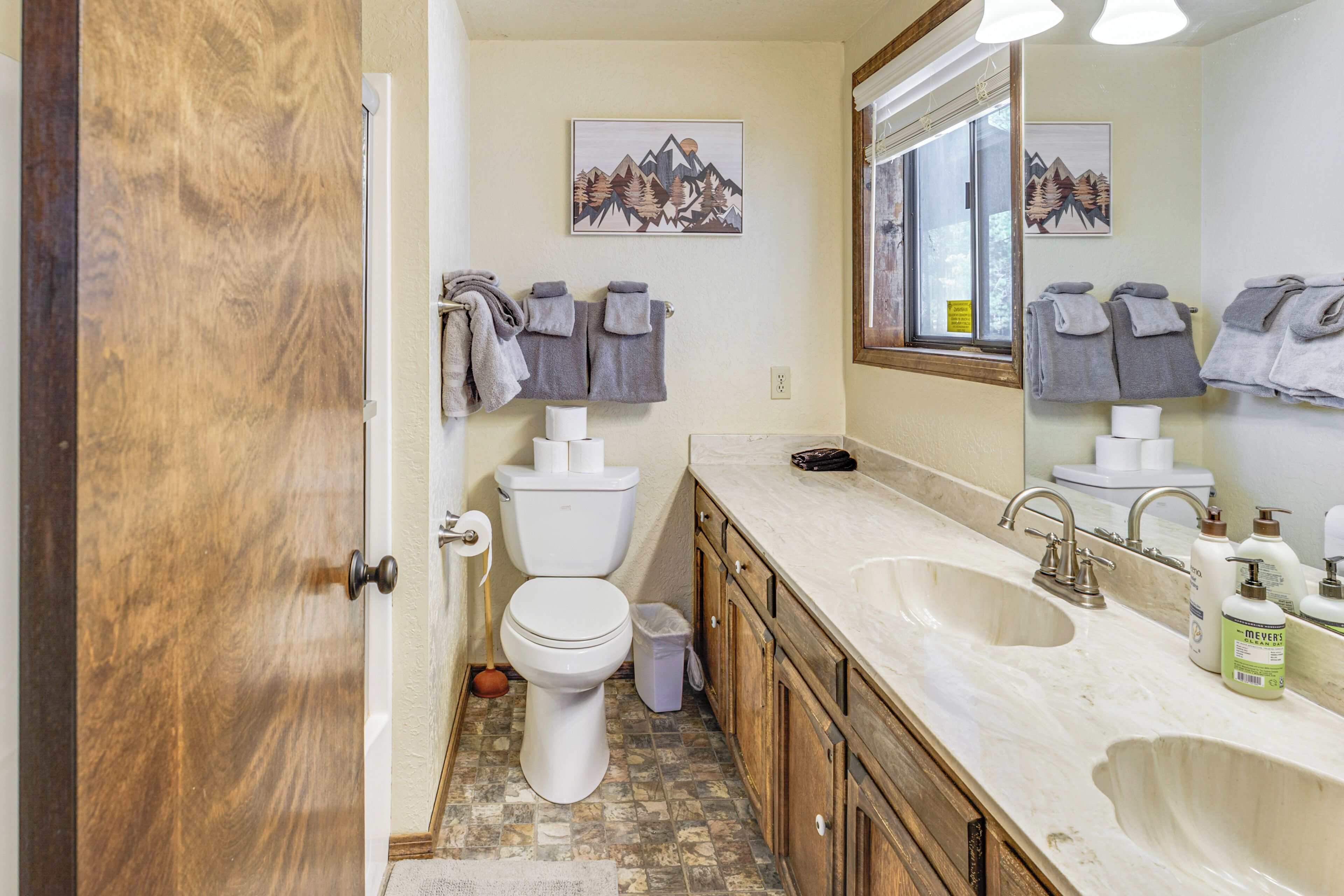 Une salle de bain équipée de toilettes, d’un lavabo et de meubles en bois. Des serviettes sont accrochées au mur et il y a une poubelle.