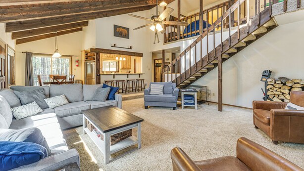 A spacious living area with a large sofa, a coffee table, and a fireplace. A staircase leads to an upper level with a balcony. - Walk to Lake & Trails: Angel Fire Home w/ Deck! (Angel Fire)