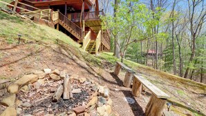 Una cabaña con terraza de madera y escaleras que llevan a un balcón, rodeada de un área boscosa con un pozo de fuego y bancos.