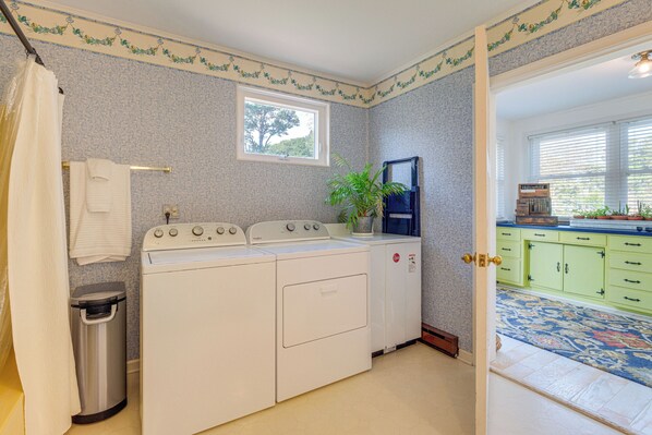 A laundry area with a washing machine and dryer, a trash can, and a window with a view of greenery. - Hot Tub & Dock: Riverfront Cottage in Deltaville (Deltaville)
