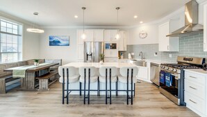 A modern kitchen with white cabinetry, a central island with bar stools, stainless steel appliances, and a large window providing natural light.