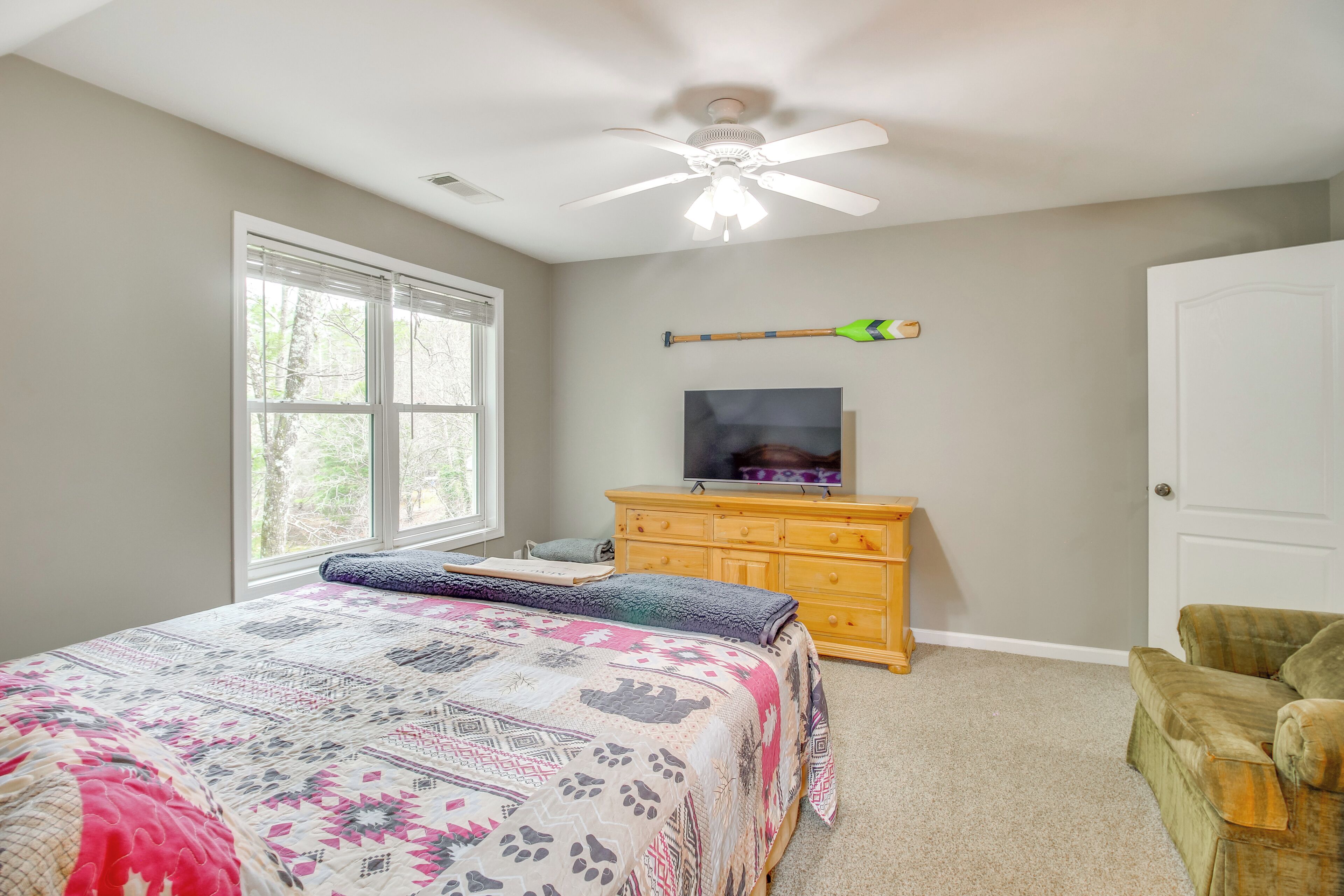 A bedroom with a bed, a ceiling fan, a TV on a wooden dresser, and a window with blinds.