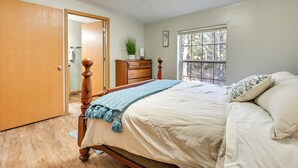 Un dormitorio con cama de madera, un tocador, una ventana con vista a árboles y una puerta que da a otra habitación.