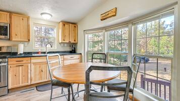 Una cocina con armarios de madera, un refrigerador de acero inoxidable, una mesa de comedor de madera con sillas de metal y ventanas amplias que ofrecen vistas a árboles y una terraza.