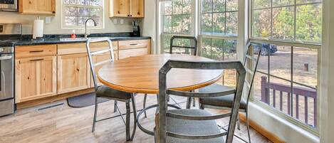 Una cocina con armarios de madera, un refrigerador de acero inoxidable, una mesa de comedor de madera con sillas de metal y ventanas amplias que ofrecen vistas a árboles y una terraza.
