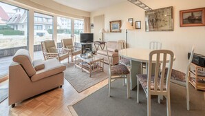 A spacious living room with a sofa, chairs, a coffee table, and a TV. Large windows offer a view of the beach and buildings outside. - Seaside Villa Apartment (Knokke-Heist)