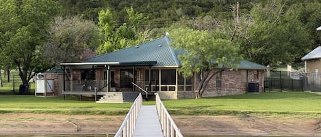 A wooden dock leading to a house with a covered porch and a stone building in the background.