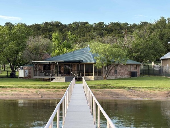 A wooden dock leading to a house with a covered porch and a stone building in the background.