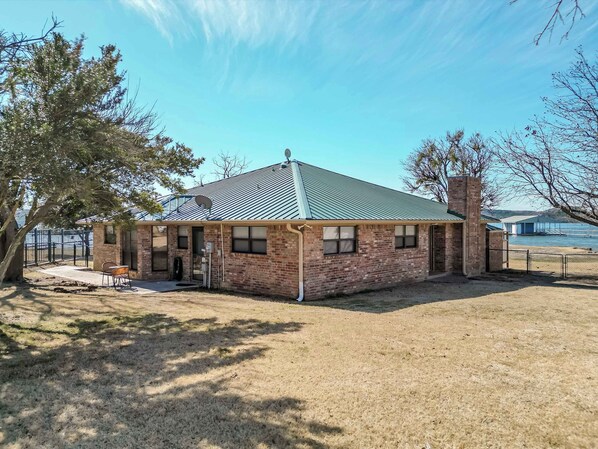 A single-story brick building with a metal roof, a satellite dish, and a fenced yard. - Family Lakefront Home with Large Porch (Graham)