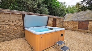 A wooden hot tub with a step for access, set on a gravel surface, surrounded by stone walls and a wooden building in the background. - Ttich Manor Retreat (Milton Keynes)