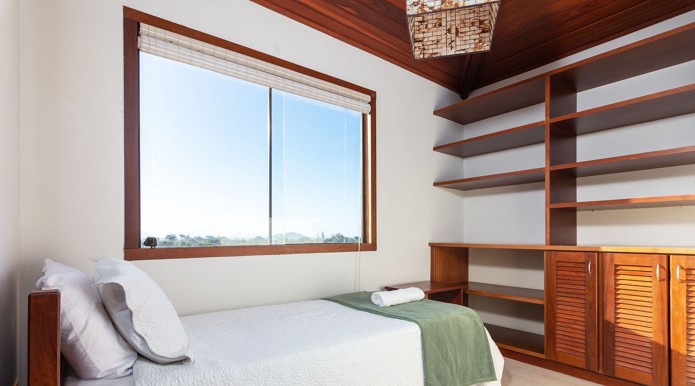 A room with a bed, a window with blinds, a wooden ceiling, and built-in shelves.