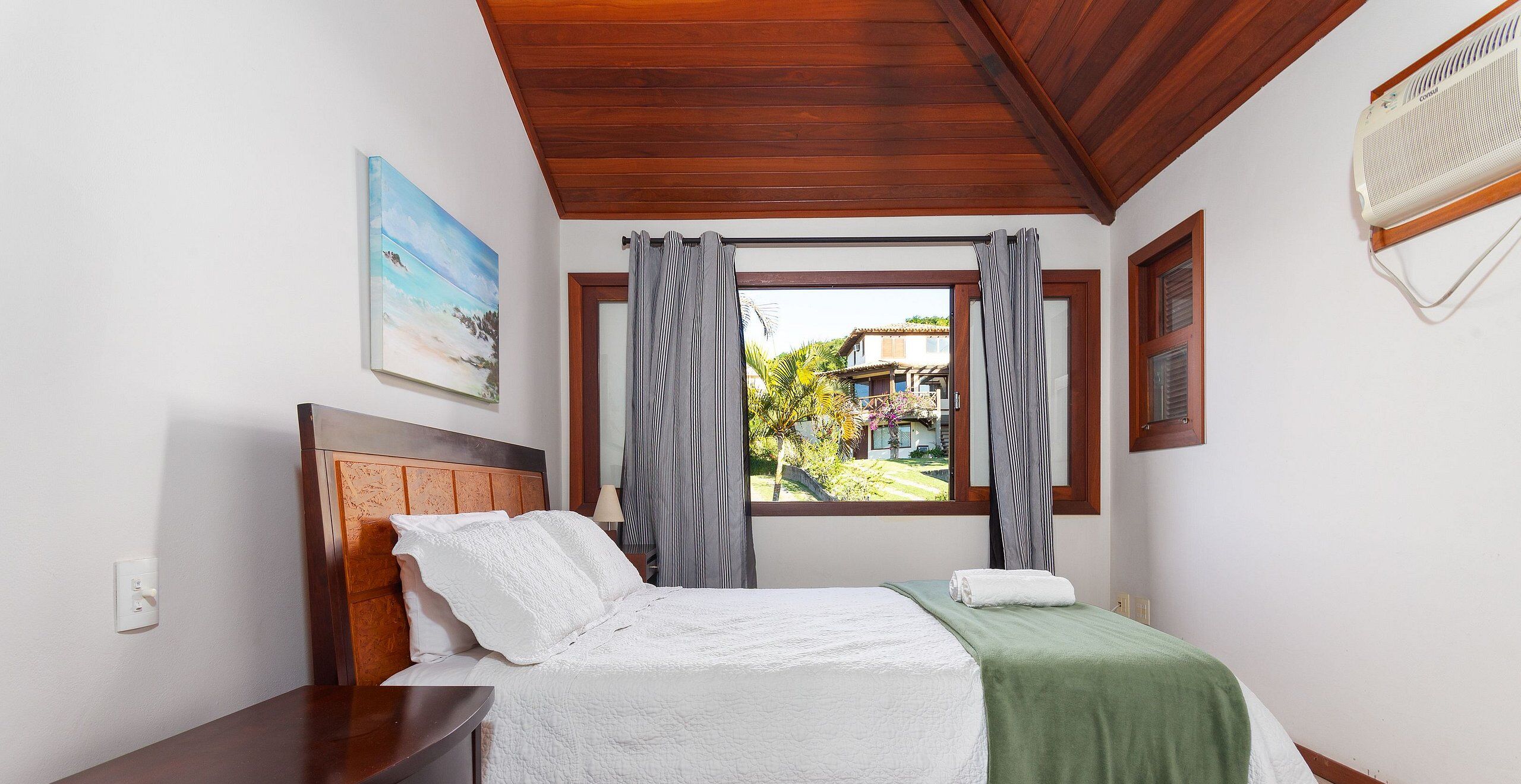 Een slaapkamer met een houten plafond, een bed met wit beddengoed, een groene plaid en een raam met gordijnen.