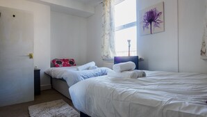 A bedroom with two beds, a nightstand, a window with curtains, and a wall-mounted artwork. - WoodHus Lodge  Cosy Large Family  Room 4 (Stoke-on-Trent)