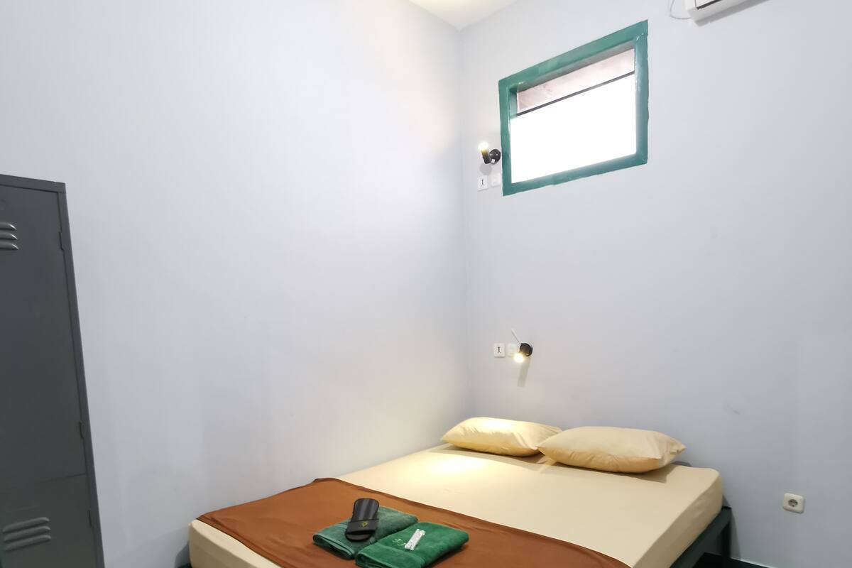 Una habitación pequeña con una cama individual, cubrecama marrón, toalla verde y un objeto negro sobre la cama. Se observa una ventana con marco verde, un aire acondicionado instalado en la pared y una puerta a la izquierda.