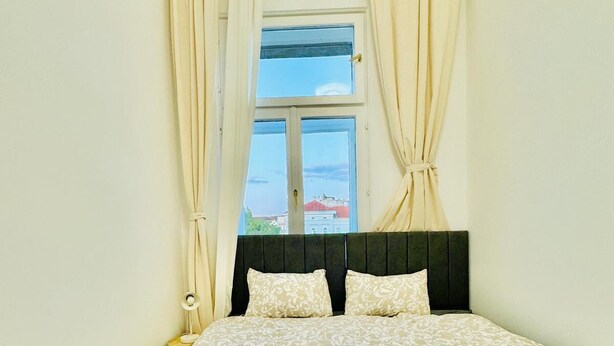A bedroom with a bed, curtains, a window, and a view of buildings. - Stylish 4BR Apt Near Westbahnhof (Vienna)
