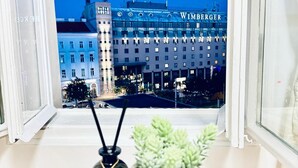 A view of a building with the name 'WIMBERGER' across the facade, a window with a view of the building, and a small potted plant on a table in the foreground. - Stylish 4BR Apt Near Westbahnhof (Vienna)