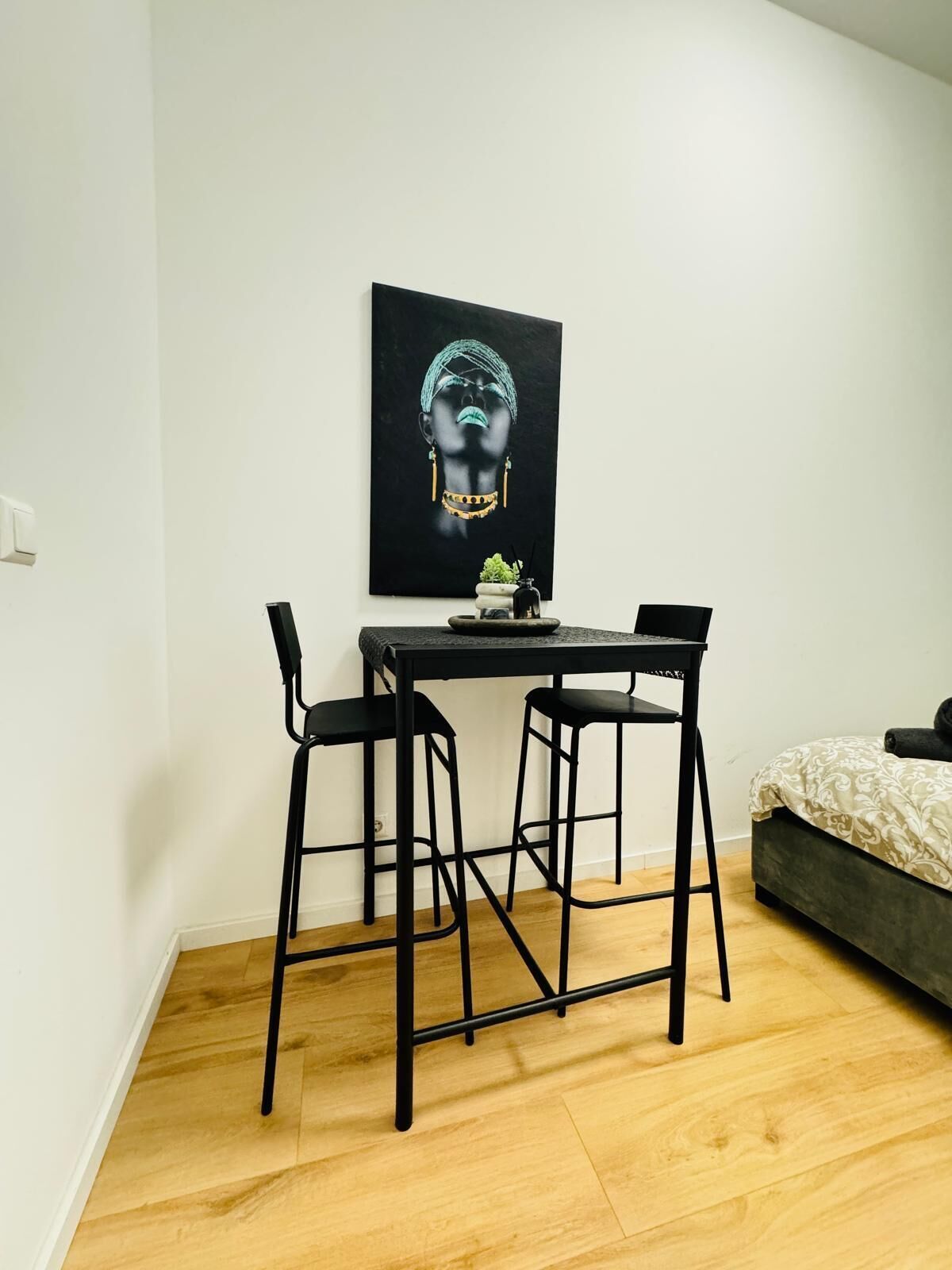 Una habitación pequeña y moderna con piso de madera, una cama con colcha estampada, una mesa negra con dos sillas y una obra de arte negra en la pared.