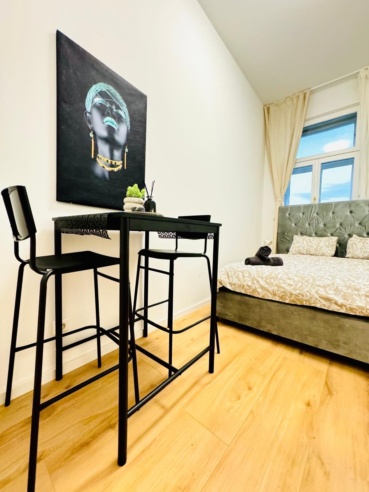 A bedroom with a bed, a desk with two chairs, a painting on the wall, and a window with curtains.
