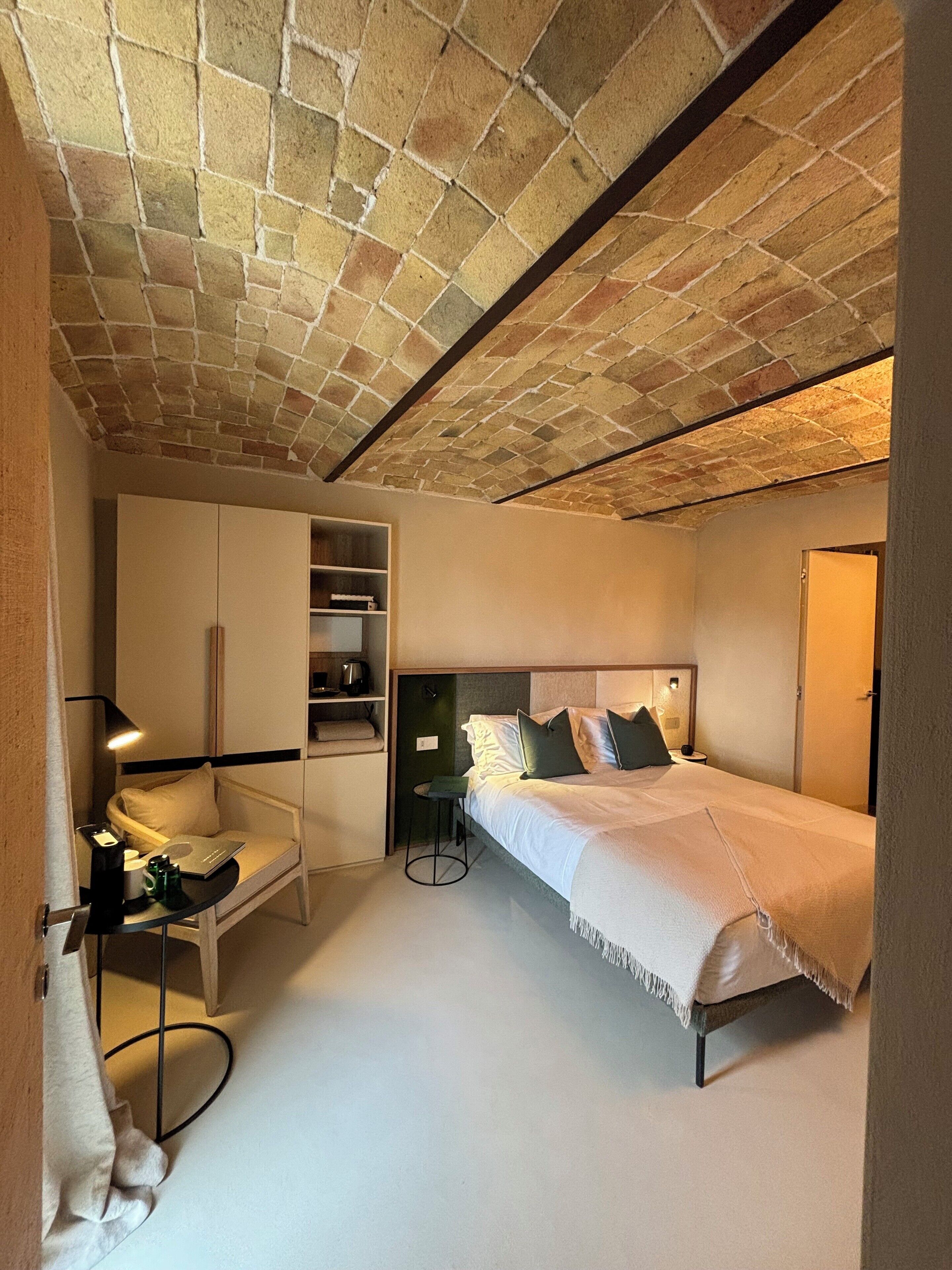 Una camera compatta con soffitto in pietra, un letto con cuscini, una sedia, una scrivania con lampada e un armadio.